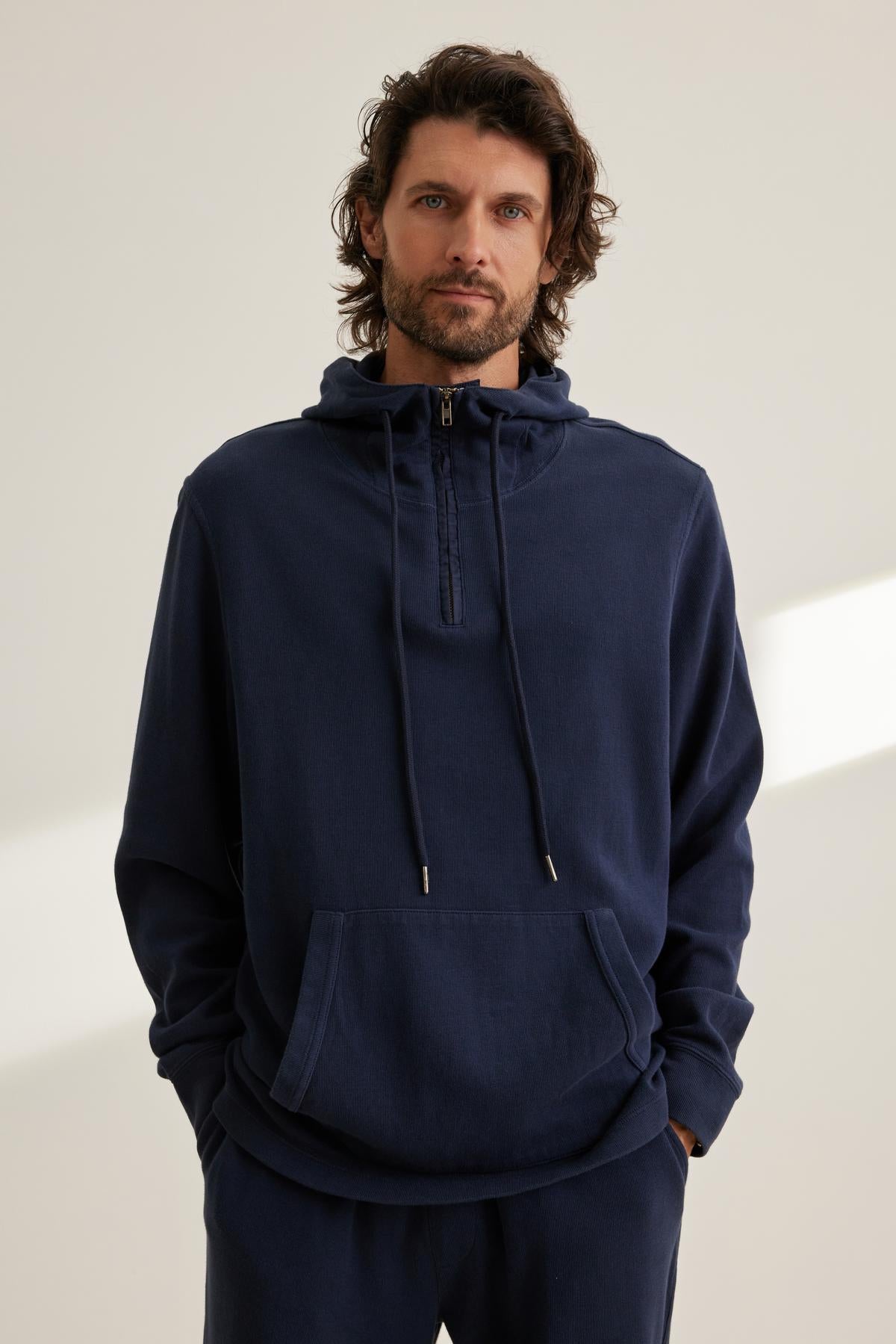   A person wearing the MORTY HOODIE, a dark blue half-zip hoodie with a front pocket from Velvet by Graham & Spencer, stands against a plain background. 