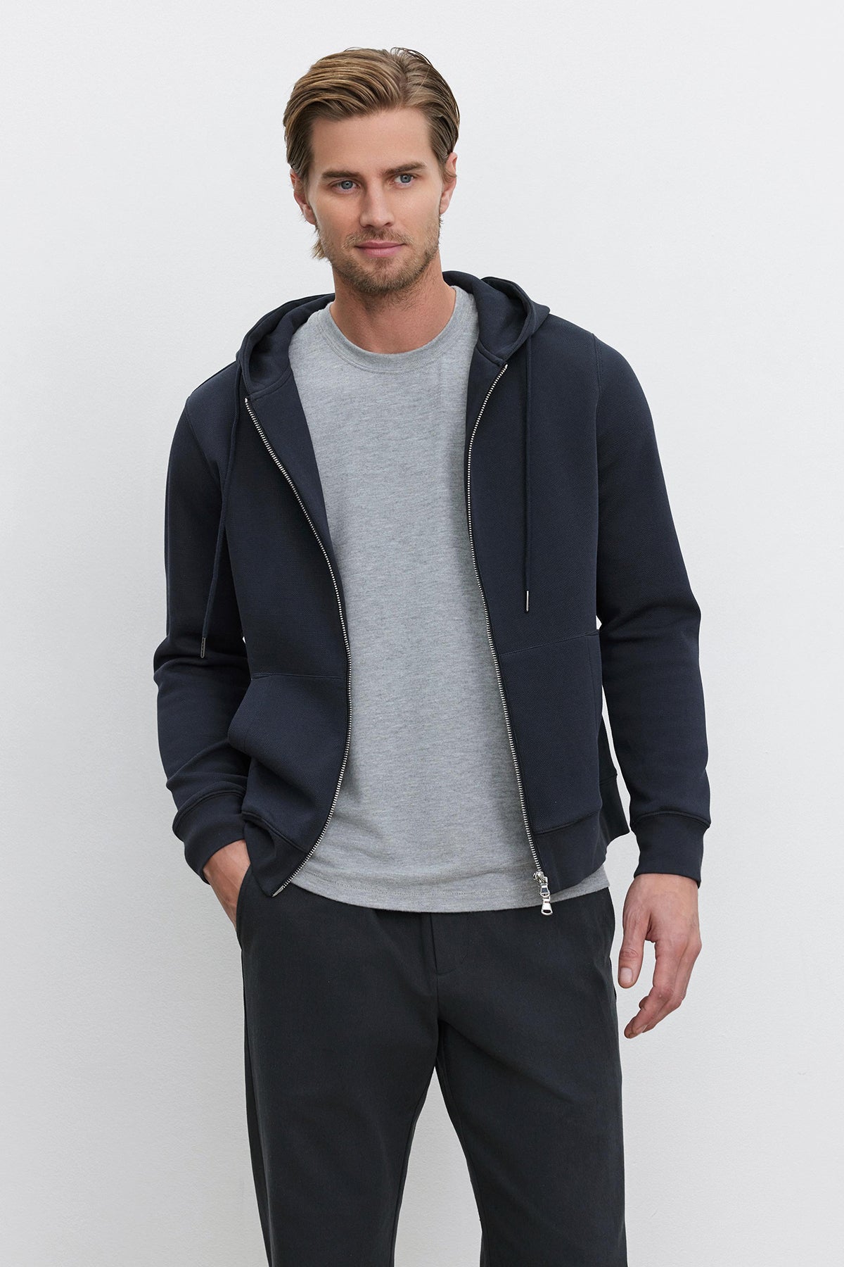   A man stands against a plain background, wearing a grey t-shirt and the LAIRD HOODIE from Velvet by Graham & Spencer, made from a textured cotton blend. His dark pants complement the look, and his left hand rests in his pocket as he looks directly at the camera. 