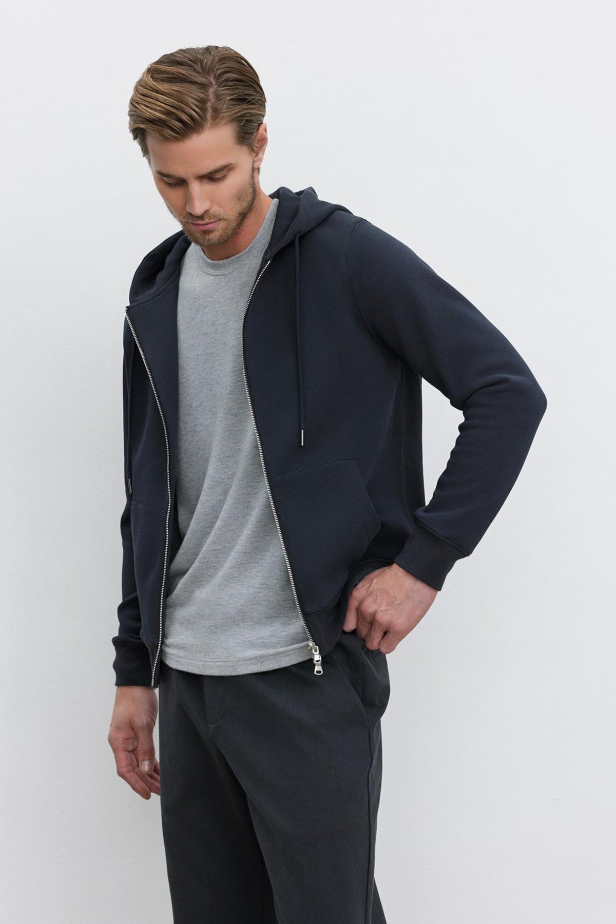   A man with short hair wears a gray T-shirt under the LAIRD HOODIE by Velvet by Graham & Spencer, made of a textured cotton blend in dark navy, paired with black pants. He stands against a white background, looking down and placing one hand in his pocket. 