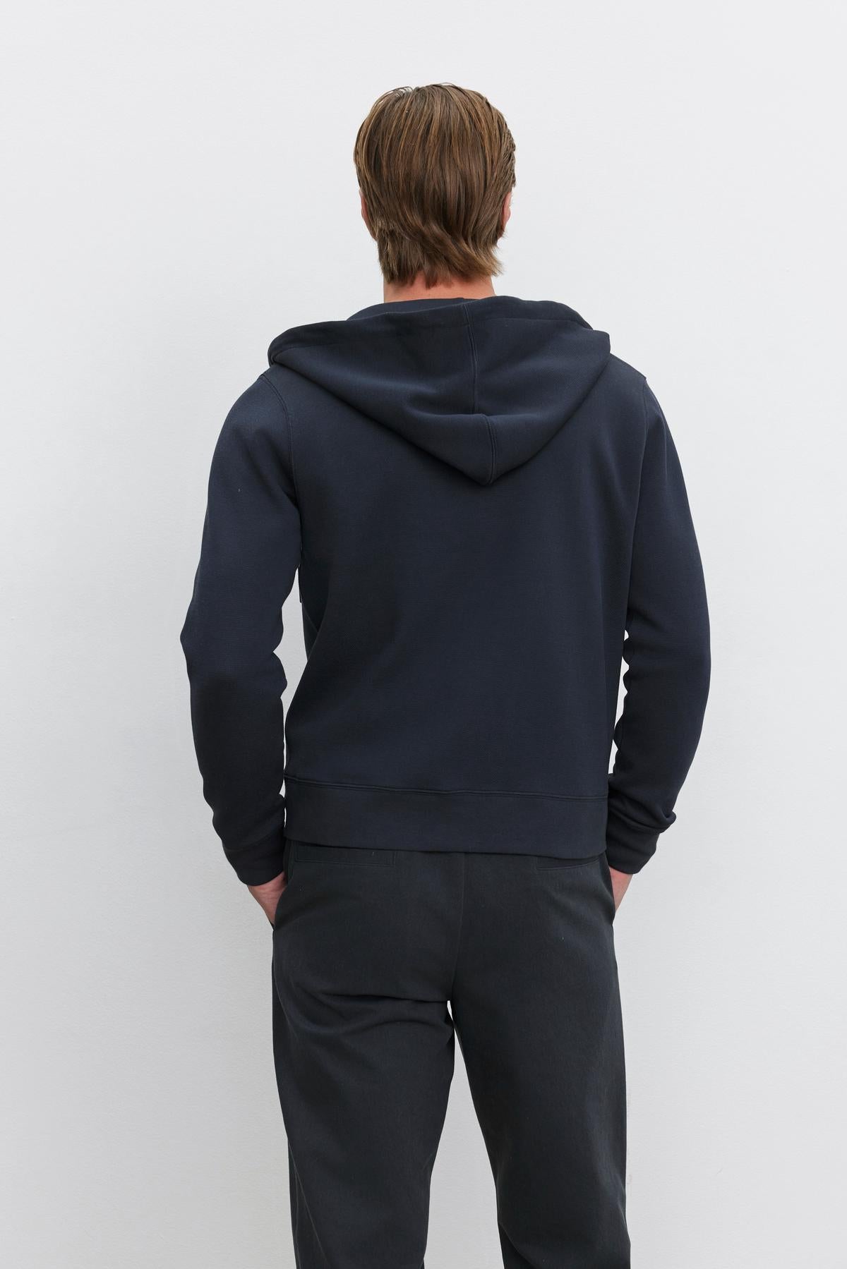   A person with light brown hair is standing and facing away from the camera, wearing the LAIRD HOODIE made of a textured cotton blend by Velvet by Graham & Spencer, along with dark pants, against a plain white background. 