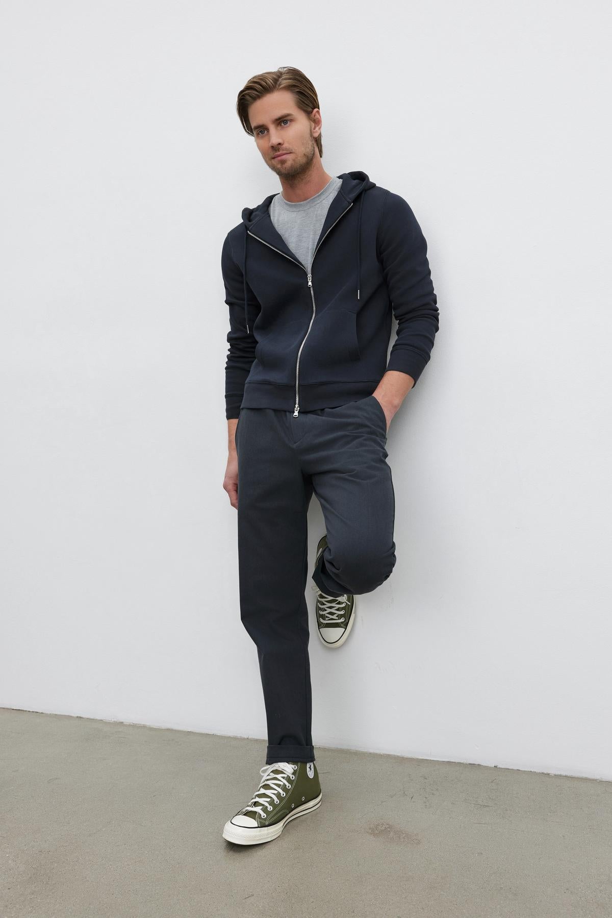   A man stands against a white wall wearing the LAIRD HOODIE by Velvet by Graham & Spencer, accompanied by a gray t-shirt, dark gray pants, and olive green Converse sneakers. His outfit showcases a stylish textured cotton blend that adds an extra layer of comfort. 