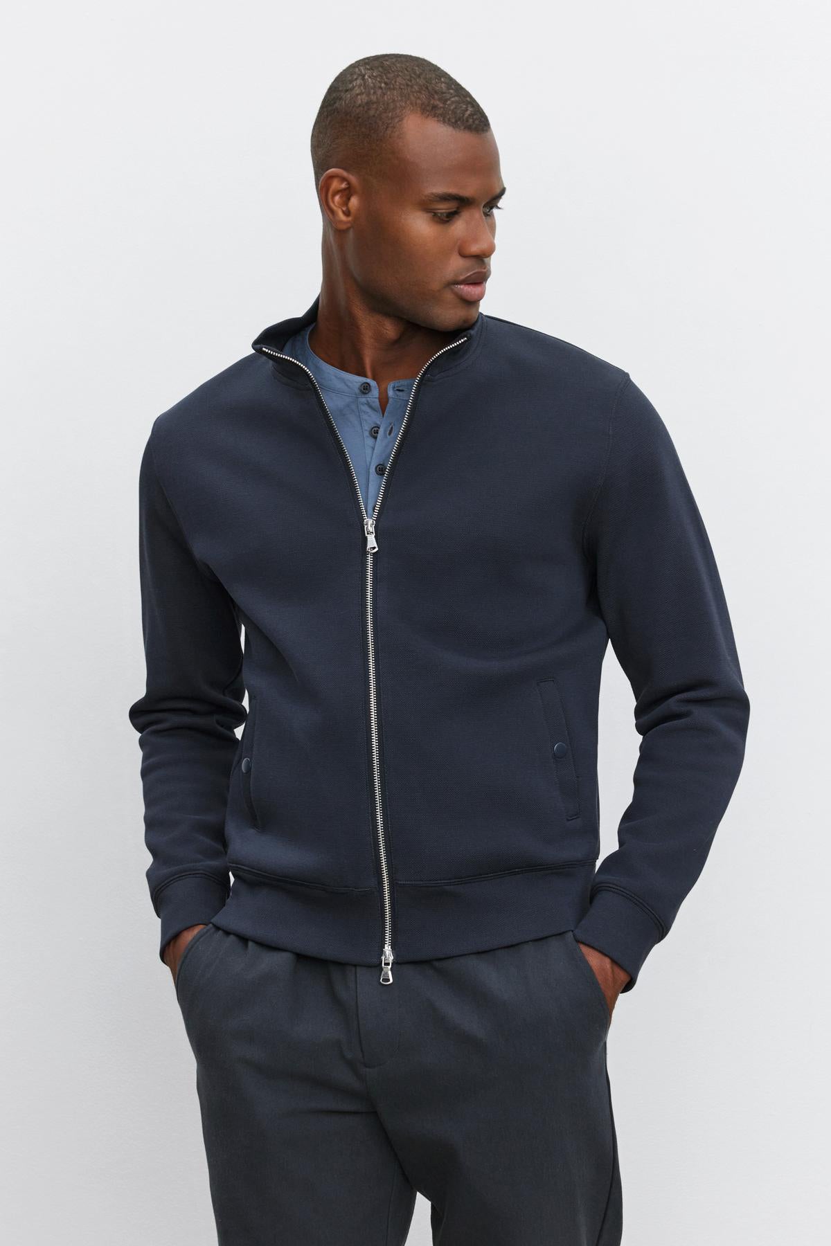   A person stands against a light background wearing the RYDELL JACKET by Velvet by Graham & Spencer, featuring a navy zip-up design with a mock neck, styled over a blue henley shirt and dark trousers, hands in pockets. 