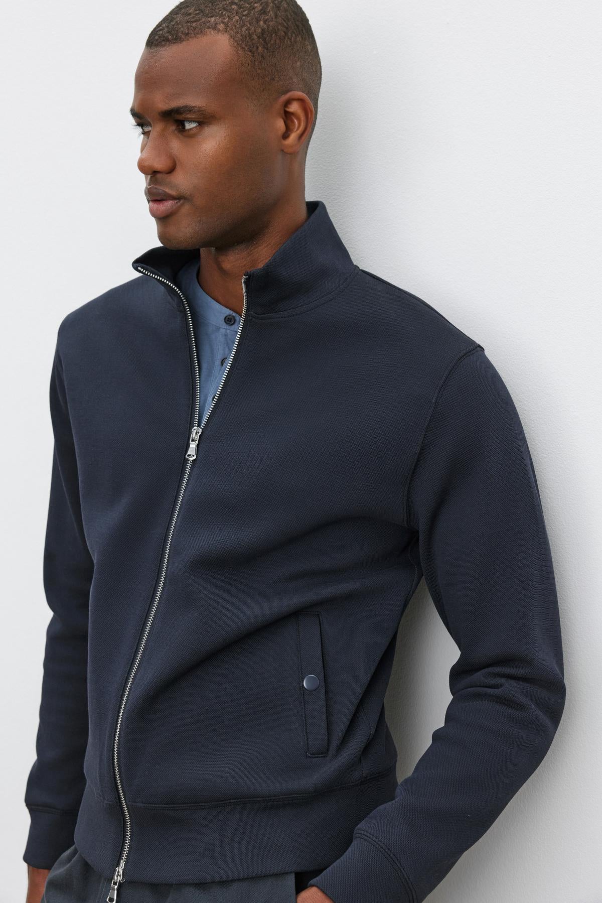   A man standing against a white wall, wearing the Velvet by Graham & Spencer RYDELL JACKET, a dark, textured cotton blend zip-up jacket, over a blue mock neck shirt, looking to his left. 