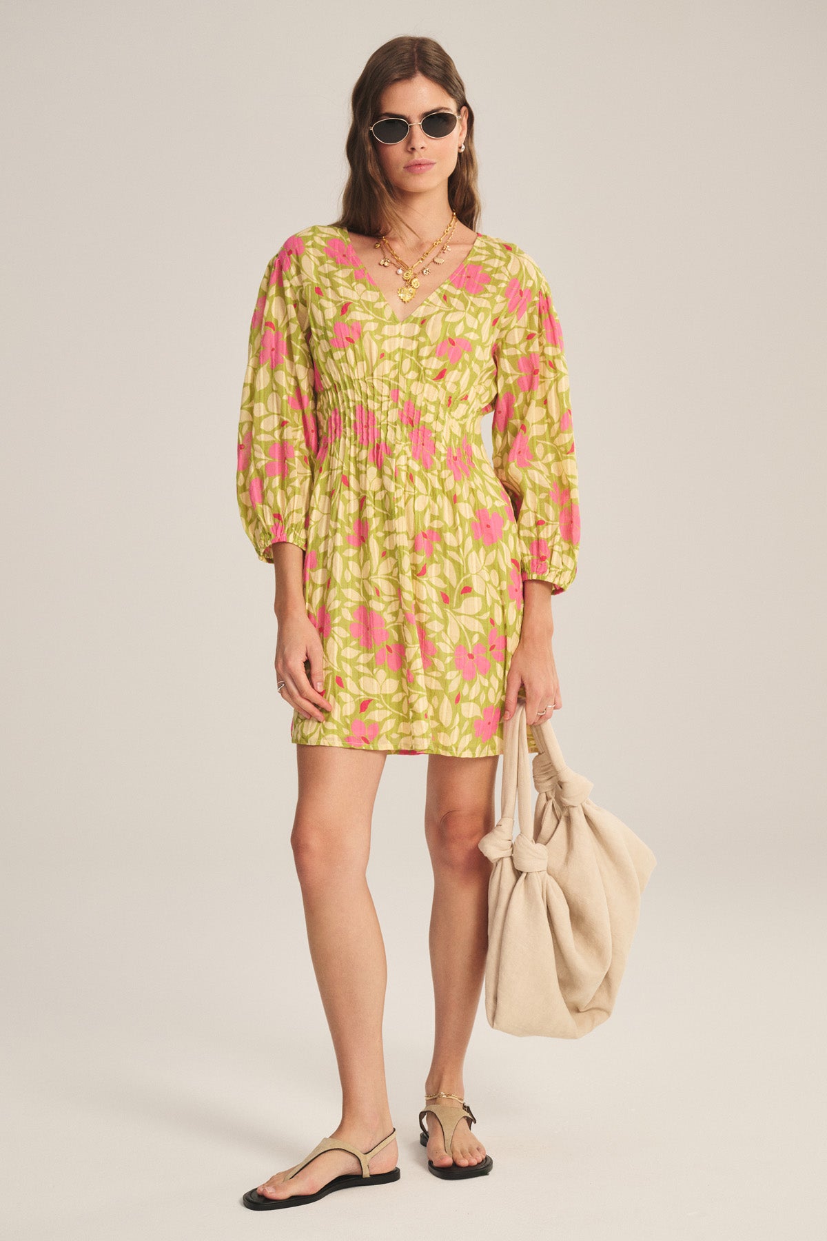 A woman in sunglasses wears the Velvet by Graham & Spencer DRAYA DRESS, a yellow and pink patterned cotton mini with puff sleeves and pleats, paired with black sandals and a large beige tote bag.