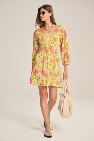 A woman in the DRAYA DRESS by Velvet by Graham & Spencer, a floral-patterned cotton mini dress with subtle Lurex detail, wears sunglasses and sandals, holding a beige bag against a plain light background.