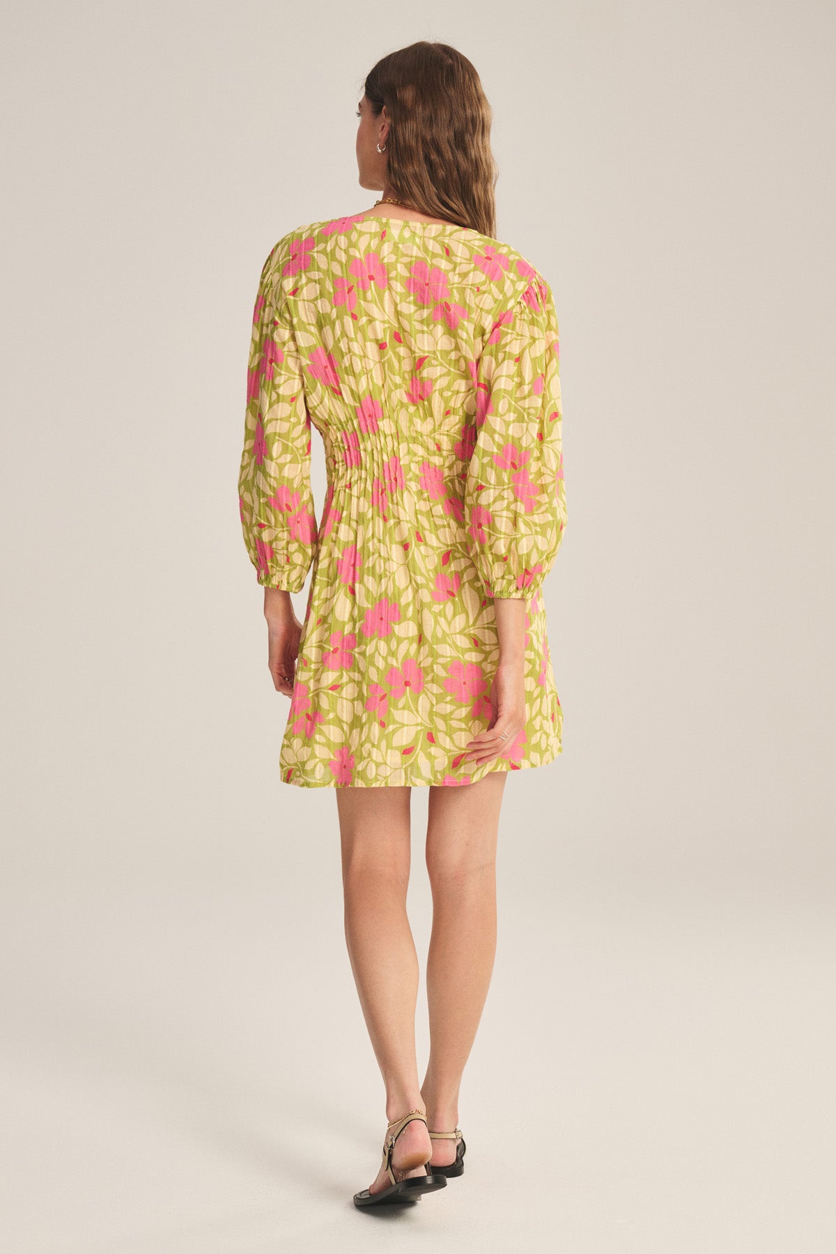 A woman faces away, wearing the DRAYA DRESS by Velvet by Graham & Spencer—a long-sleeve yellow cotton dress with a pink and white floral pattern—paired with black sandals against a plain background.