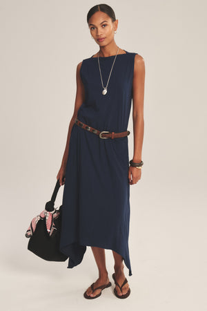 Woman wearing the DREE DRESS by Velvet by Graham & Spencer, a sleeveless navy style with a handkerchief hem and brown belt, paired with sandals, jewelry, and a black bag adorned with a pink scarf.