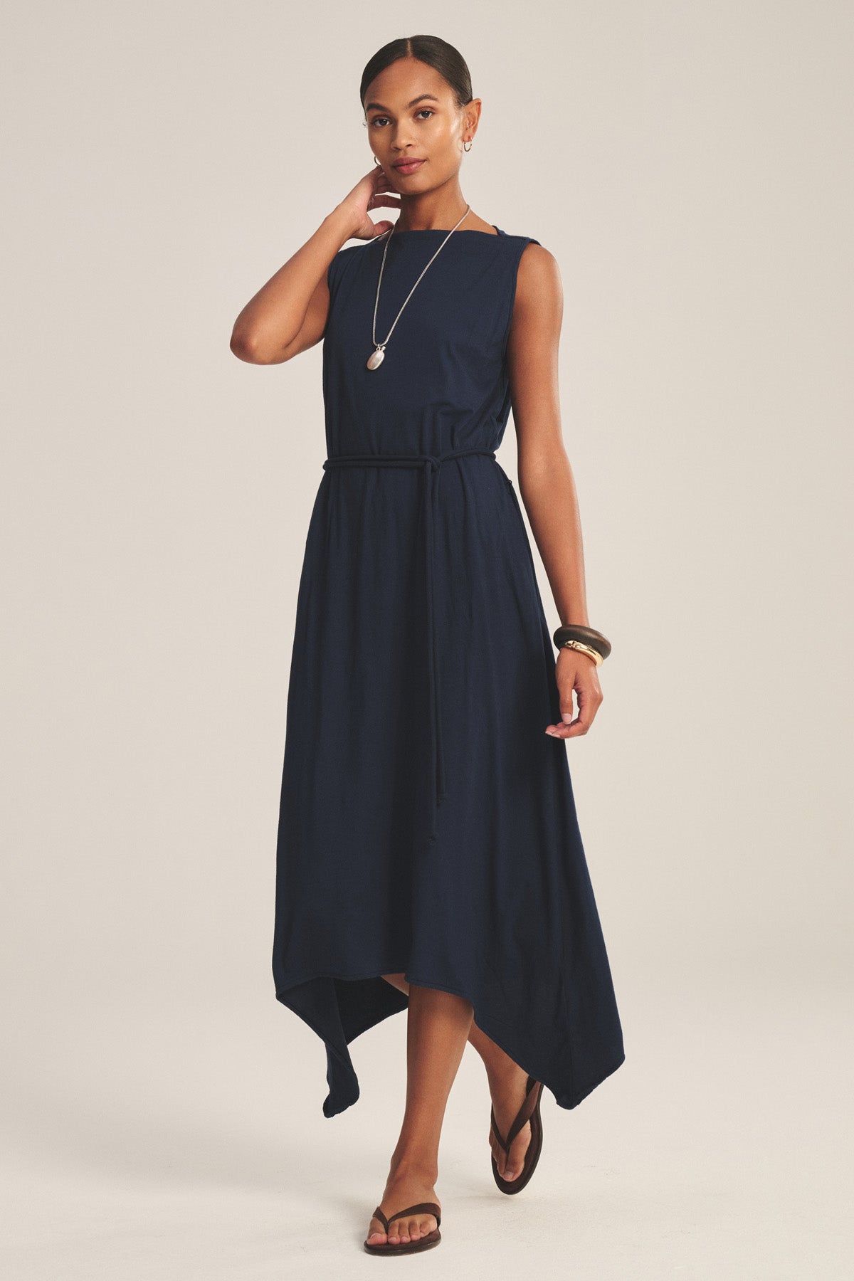 A woman models the Velvet by Graham & Spencer DREE DRESS, a sleeveless navy blue dress with a handkerchief hem, styled with a black belt, necklace, bracelet, and brown sandals against a plain background.-41008618635457