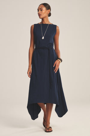 A woman models the DREE DRESS by Velvet by Graham & Spencer—a sleeveless navy blue handkerchief hem dress—styled with a thin belt, long necklace, bangles, and black sandals against a plain light background.