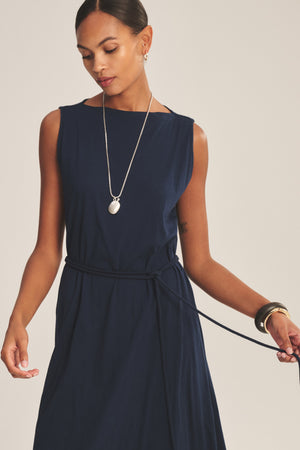 A woman in the DREE DRESS by Velvet by Graham & Spencer, featuring a thin waist tie, accessorizes with a long necklace and bracelets, her hair tied back as she looks down.