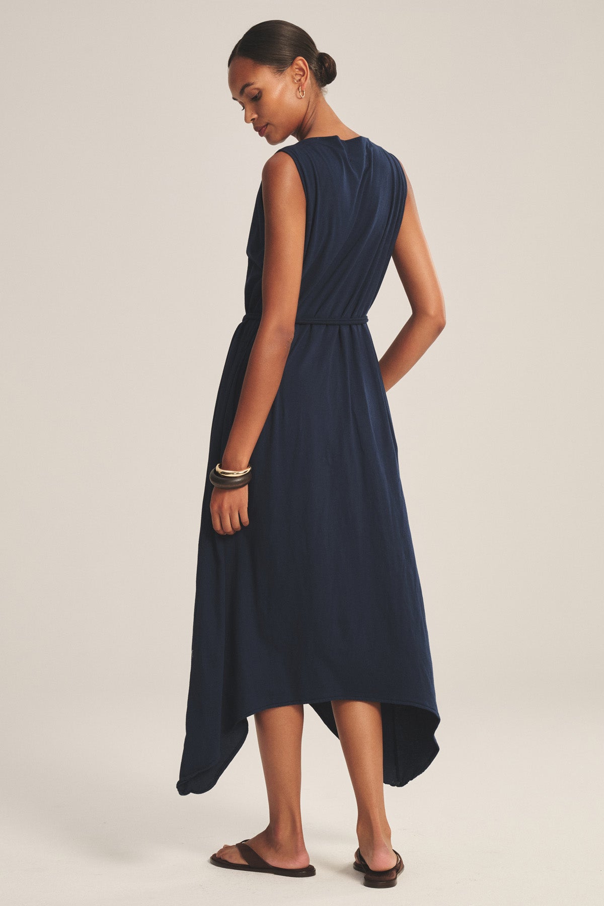 A woman stands with her back to the camera in the DREE DRESS by Velvet by Graham & Spencer—a sleeveless navy blue dress with a handkerchief hem—paired with sandals against a plain background.-41008618668225