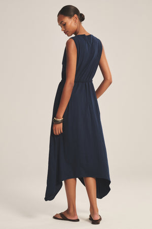A woman stands with her back to the camera in the DREE DRESS by Velvet by Graham & Spencer—a sleeveless navy blue dress with a handkerchief hem—paired with sandals against a plain background.