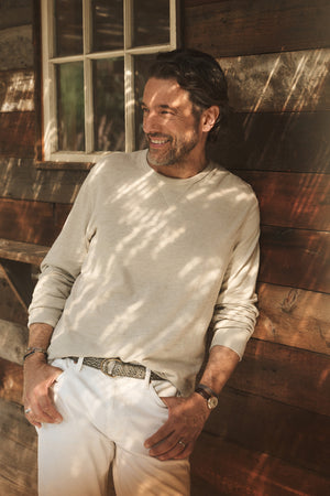 Wearing the Velvet by Graham & Spencer DUSTIN TEE in a relaxed fit with white pants, a man stands smiling against a wooden wall as sunlight and shadows play across him.