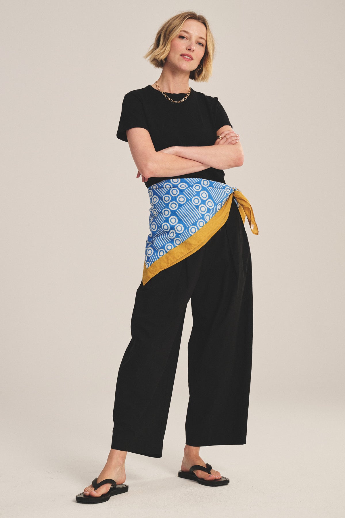 A person stands in a relaxed pose wearing a black t-shirt, EASTON PANT by Velvet by Graham & Spencer—cropped cotton poplin with an elastic waist—flip-flops, and a blue and yellow patterned wrap tied at the waist.
