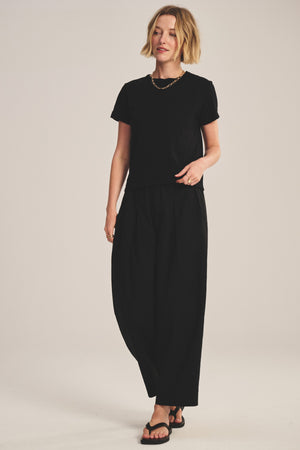 A woman stands against a plain background, wearing a black t-shirt, the EASTON PANT by Velvet by Graham & Spencer—cropped cotton poplin with an elastic waist—paired with sandals and a gold chain necklace.