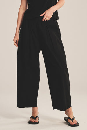 A person wears the EASTON PANT by Velvet by Graham & Spencer—loose black cotton poplin pants with an elastic waist and cropped length—paired with a black top and flip-flops, standing against a neutral background.