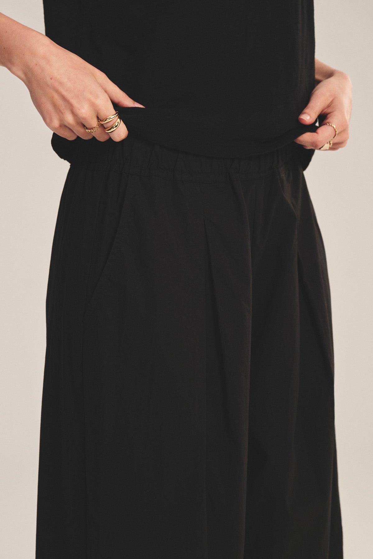 A person wearing the EASTON PANT by Velvet by Graham & Spencer—black cotton poplin with an elastic waist—pairs it with a black top, hands resting near the waistband and gold rings visible on their fingers.