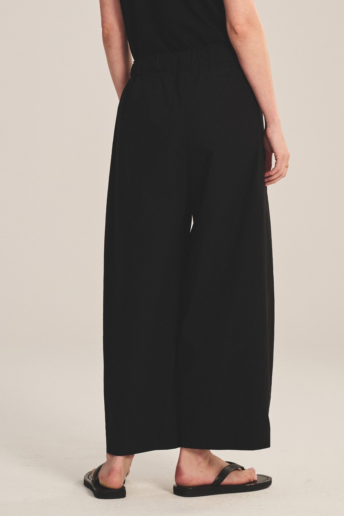 A person stands with their back to the camera in Velvet by Graham & Spencer’s EASTON PANT—loose, cropped black cotton poplin with an elastic waist—paired with a black top and sandals, set against a plain light background.-41296712794305