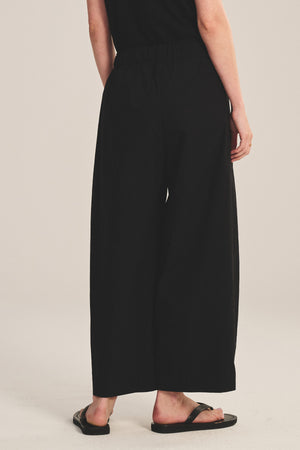 A person stands with their back to the camera in Velvet by Graham & Spencer’s EASTON PANT—loose, cropped black cotton poplin with an elastic waist—paired with a black top and sandals, set against a plain light background.