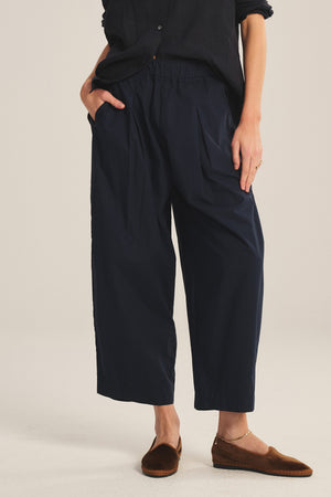 Wearing the EASTON PANT by Velvet by Graham & Spencer—loose-fitting, navy, cropped with an elastic waist—a person pairs them with a black top and brown flats, standing with one hand in their pocket.