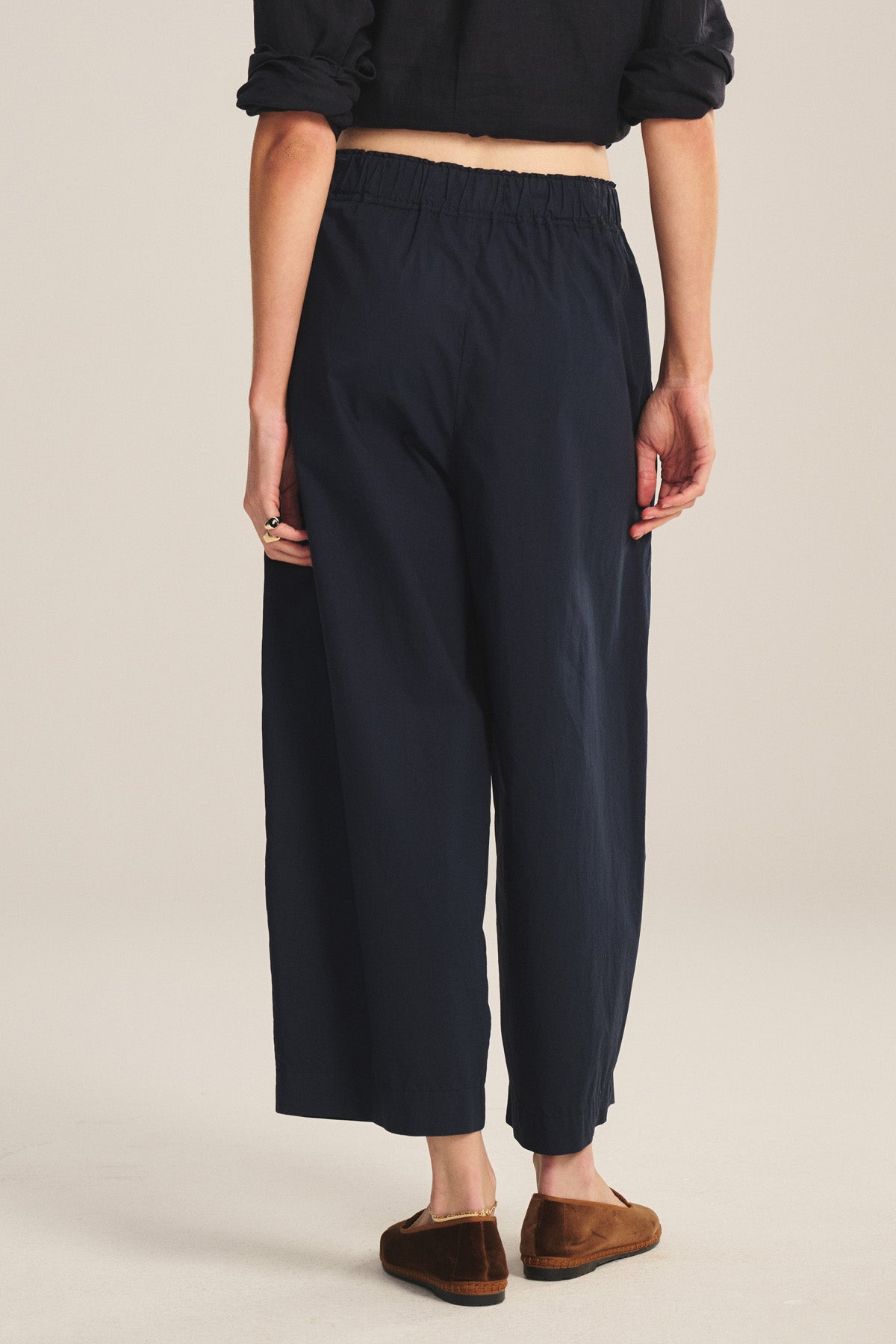 Wearing the EASTON PANT by Velvet by Graham & Spencer—loose-fitting, dark navy, cropped with an elastic waist—paired with a rolled-sleeve black top and brown shoes, they stand back to the camera against a neutral backdrop; a modern essential.
