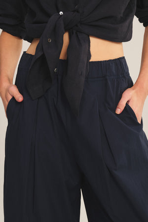 A person wearing the EASTON PANT by Velvet by Graham & Spencer—navy, loose-fit, elastic waist cropped pants styled with a black button-up shirt tied at the waist and hands in pockets for an effortlessly modern look.