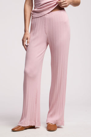 A person wearing the ALEMI POINTELLE PANT by Velvet by Graham & Spencer in light pink with subtle ribbed pointelle accents, styled with brown shoes, poses with one hand on their hip and the other gently touching the pants.