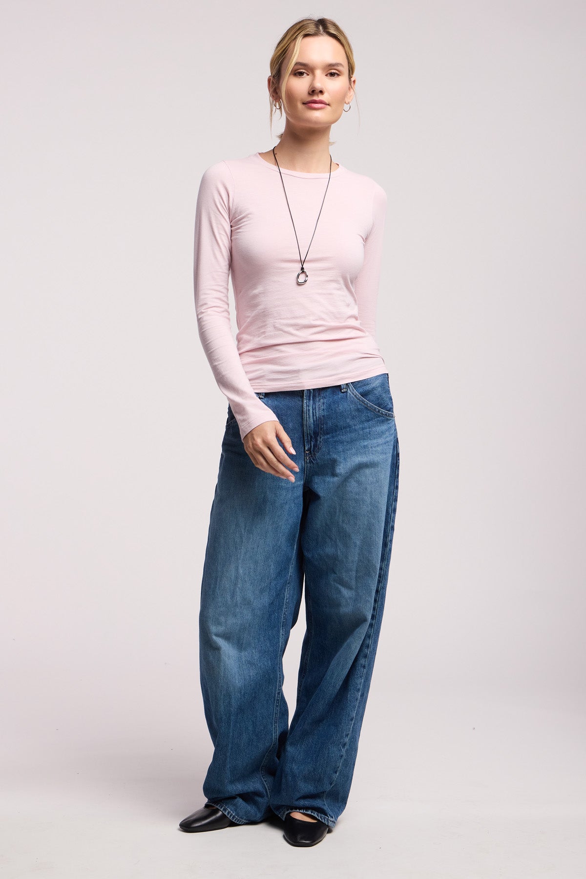 A woman faces forward in a light pink ZOFINA TEE by Velvet by Graham & Spencer, styled with loose blue jeans, black shoes, and a pendant necklace against a plain light backdrop.-40598576169153