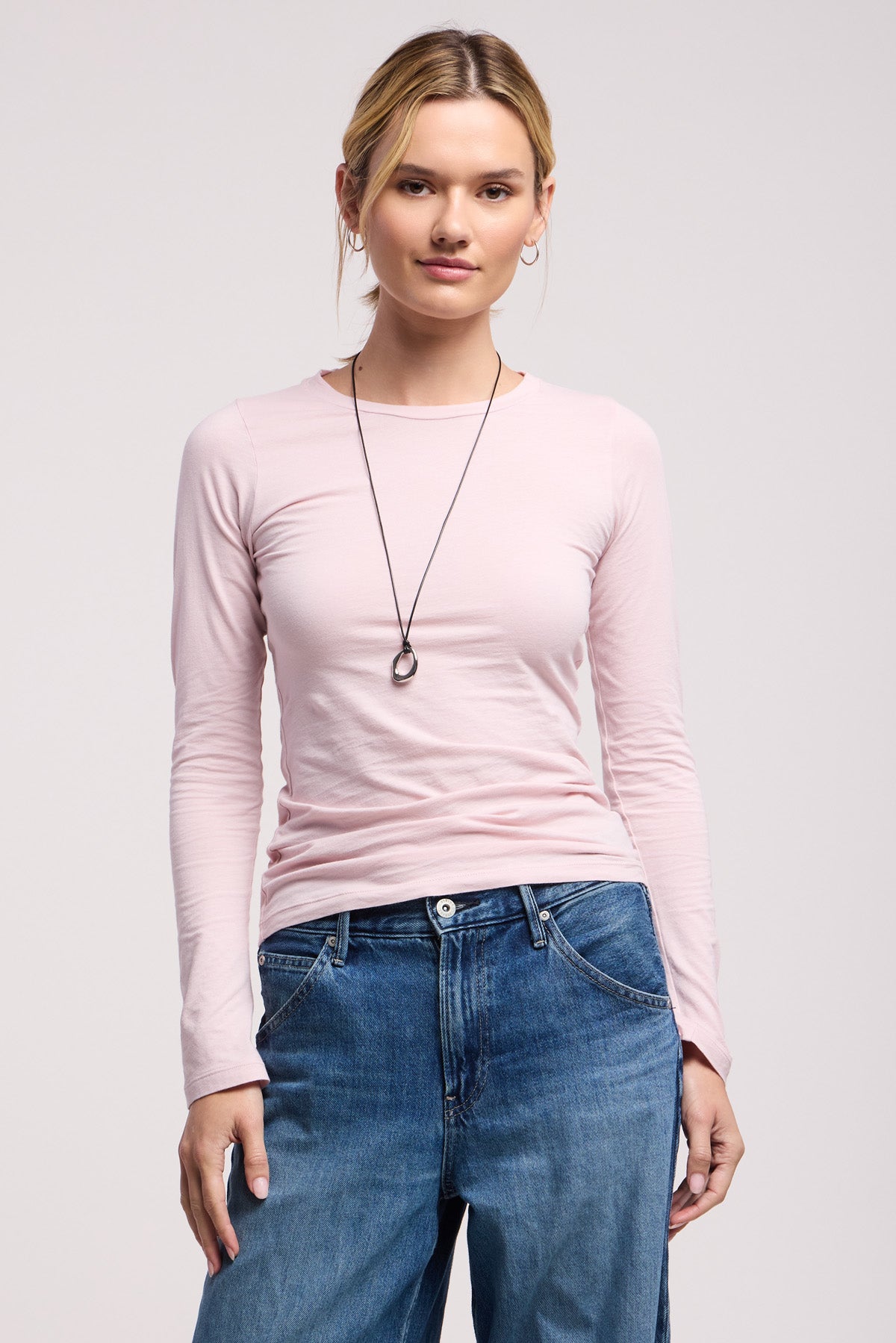 A person stands facing forward, wearing the ZOFINA TEE by Velvet by Graham & Spencer, a light pink fitted long sleeve top from the basics collection, styled with blue jeans and a circular pendant necklace.