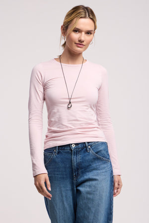 A woman stands facing the camera, wearing the Velvet by Graham & Spencer ZOFINA TEE in light pink, styled with blue jeans and a pendant necklace against a plain background.