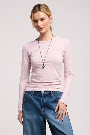 A person stands facing forward against a plain background, wearing the ZOFINA TEE by Velvet by Graham & Spencer—a light pink fitted long sleeve top—paired with blue jeans and a necklace.