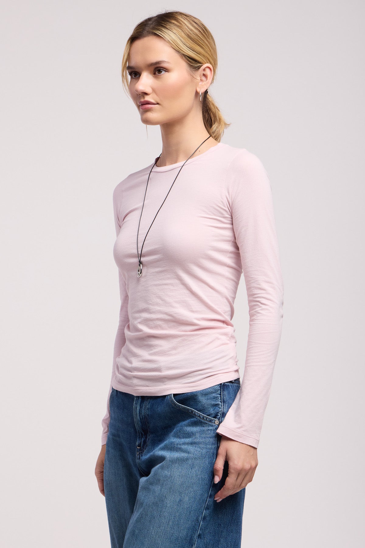 A person stands against a plain background wearing the ZOFINA TEE by Velvet by Graham & Spencer in light pink, styled with blue jeans and a delicate pendant necklace.