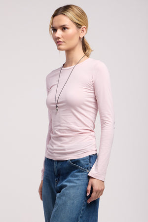 A person stands against a plain background wearing the ZOFINA TEE by Velvet by Graham & Spencer in light pink, styled with blue jeans and a delicate pendant necklace.