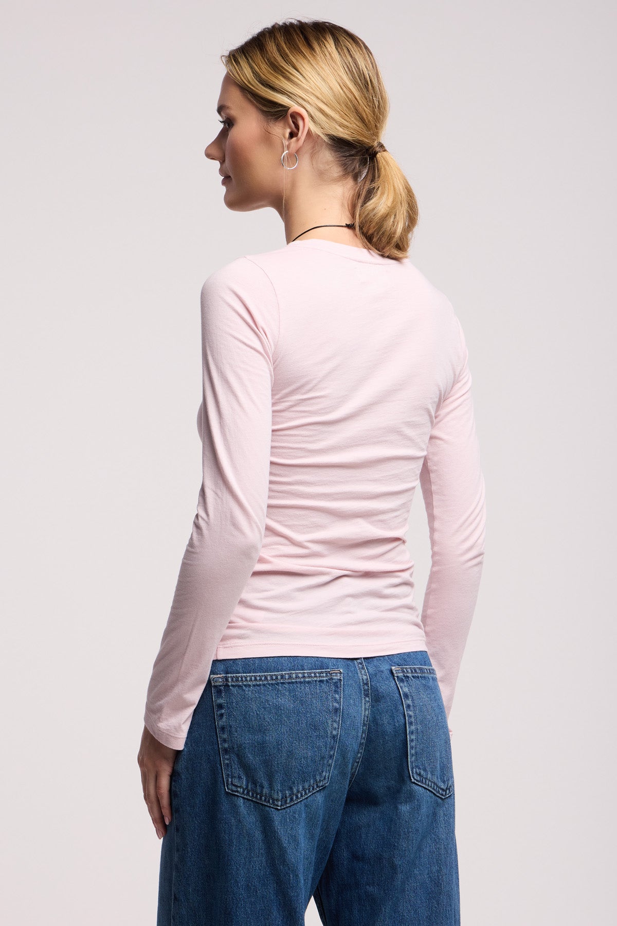 A woman stands facing away, wearing the ZOFINA TEE in light pink by Velvet by Graham & Spencer from our basics collection, paired with blue jeans.