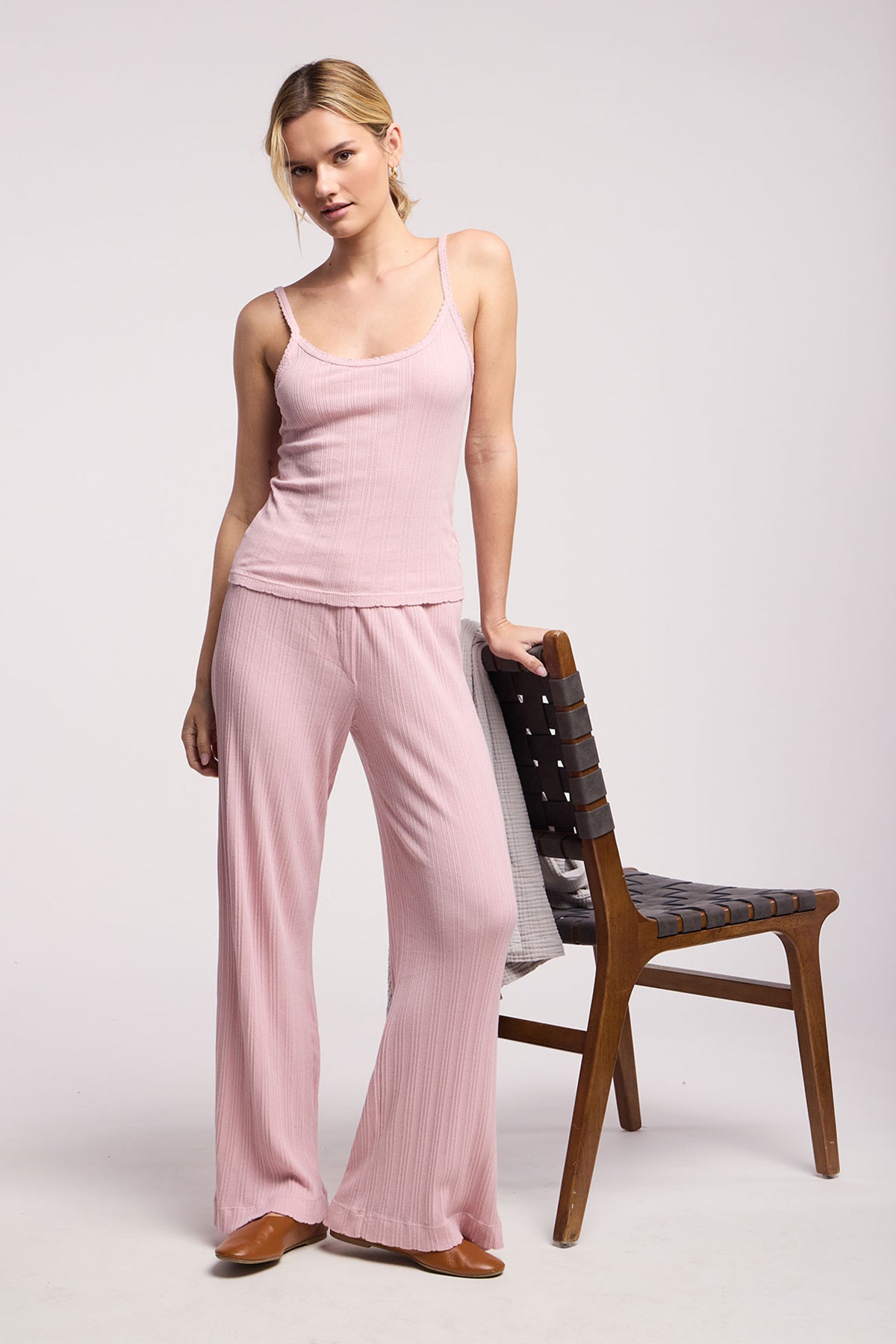 A woman in the ALEMI POINTELLE PANT by Velvet by Graham & Spencer, paired with a light pink ribbed tank top, stands next to a wooden chair, resting one hand on its backrest.-40416877510849