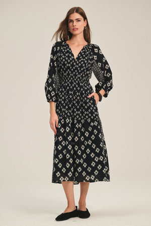 A woman wears the ELEN DRESS by Velvet by Graham & Spencer, a black midi/maxi dress with white geometric patterns and long sleeves, paired with black flats, standing against a plain light background.