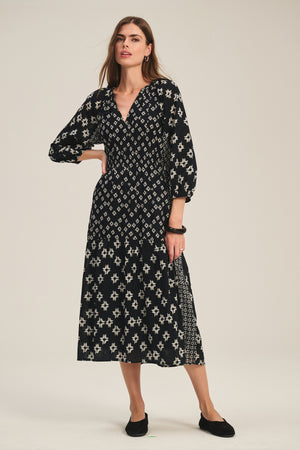 A woman wears the ELEN DRESS by Velvet by Graham & Spencer, a black cotton midi dress with white geometric patterns and long sleeves, paired with black flats against a plain light background.
