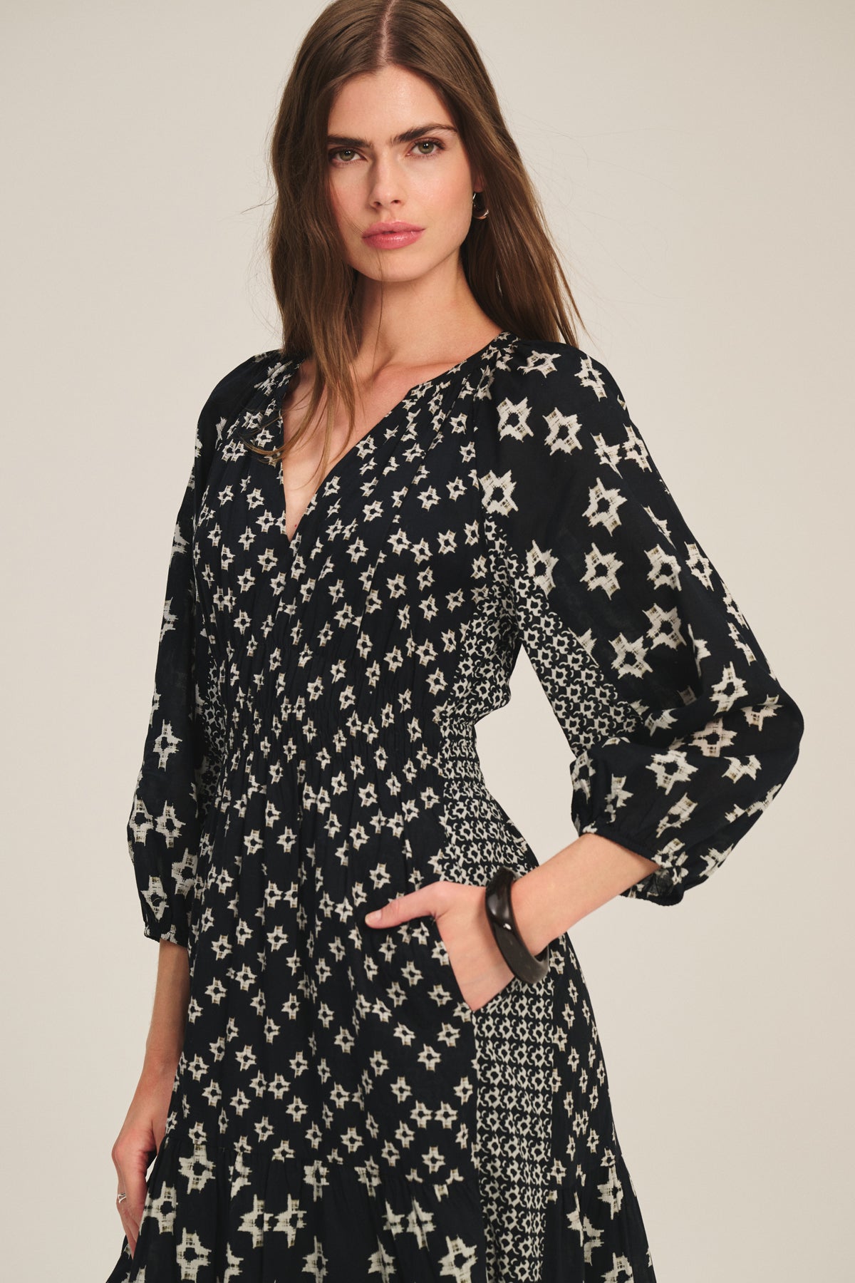   A woman wears the ELEN DRESS by Velvet by Graham & Spencer, a long-sleeved black midi maxi dress featuring a white geometric star pattern, standing with one hand in her pocket against a plain background. 