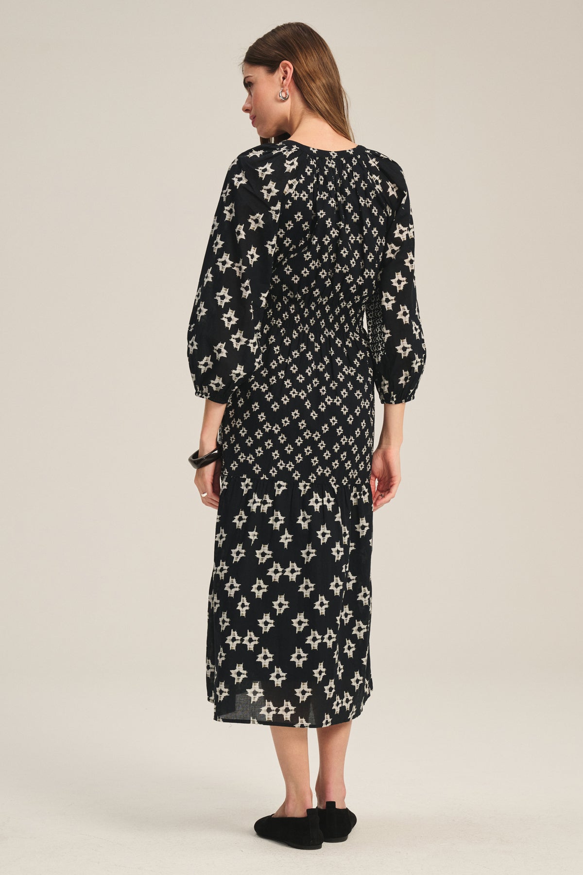 A woman with straight brown hair and black shoes faces away, wearing the ELEN DRESS by Velvet by Graham & Spencer—a black midi maxi dress with white geometric and star patterns. The background is plain and light-colored.-39815218233537