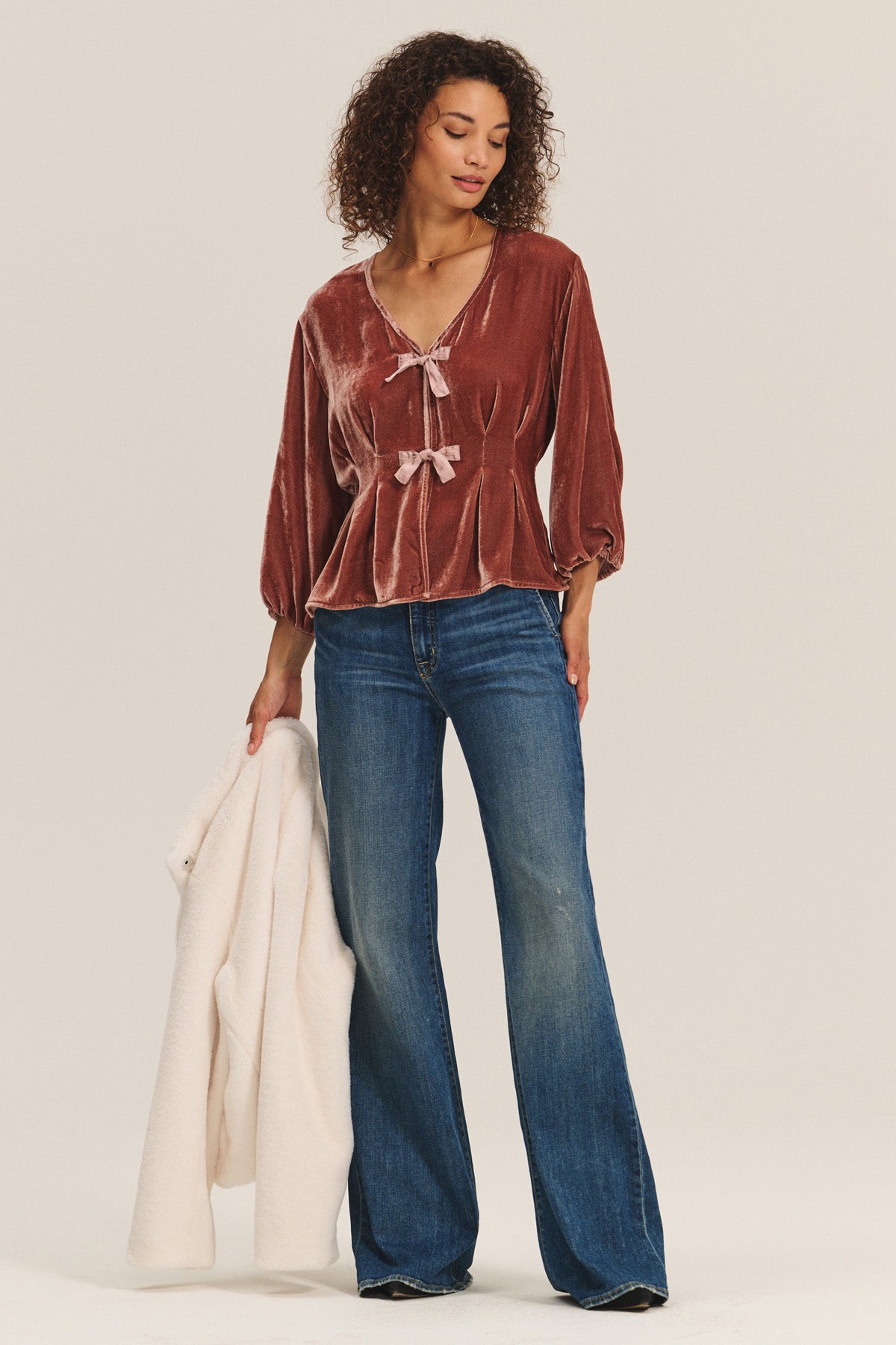Wearing the ELIDA TOP by Velvet by Graham & Spencer in rust silk velvet with bow ties and pleats, paired with blue flared jeans, a person holds a white coat in their left hand against a plain background.-40263648280769