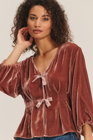 A woman with curly hair models the ELIDA TOP by Velvet by Graham & Spencer—a rust-colored silk velvet top featuring long sleeves, pleated details, and front bows—against a neutral background for a chic holiday look.
