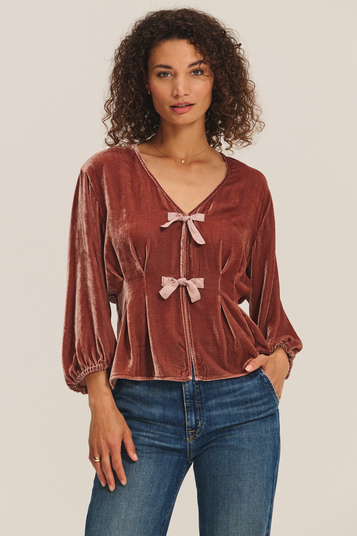 A woman wears the ELIDA TOP by Velvet by Graham & Spencer—a rose-colored silk velvet top with pleated details and front pink bows—paired with blue jeans, against a plain background for a perfect holiday look.-40263648215233