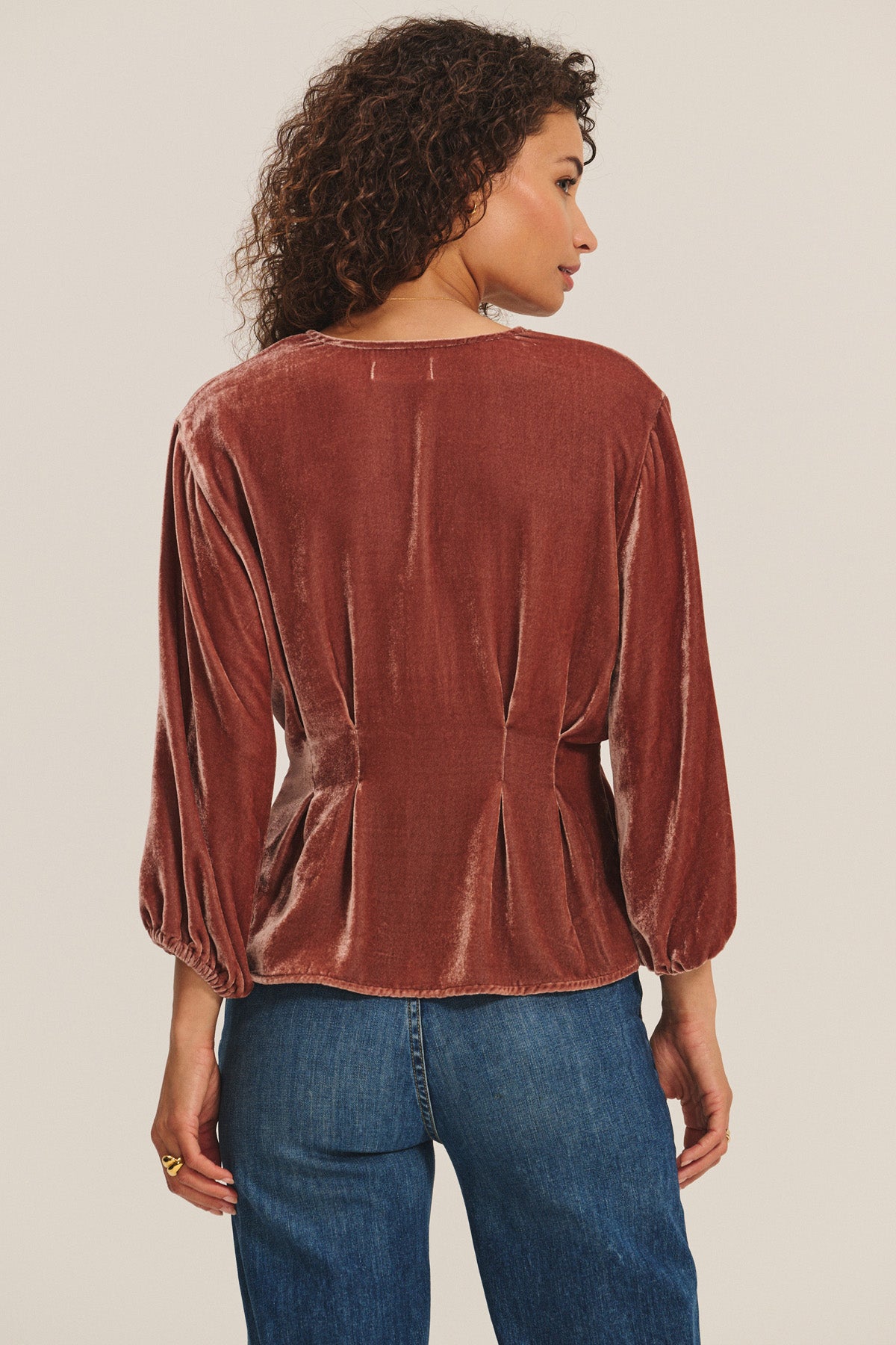 A woman stands with her back to the camera, wearing the ELIDA TOP by Velvet by Graham & Spencer—a rust-colored, long-sleeve silk velvet blouse with pleated details—styled with blue jeans for a chic holiday look.-40263648182465