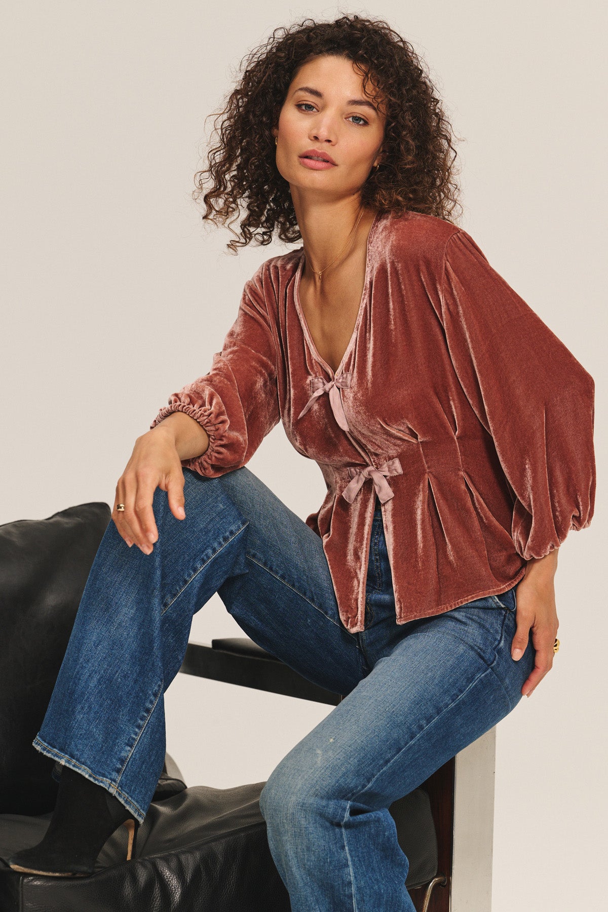 A woman sits on a chair wearing the ELIDA TOP by Velvet by Graham & Spencer, featuring wide sleeves and pleated details, paired with blue jeans. She looks at the camera with a neutral expression for a chic, understated holiday look.-40263648149697