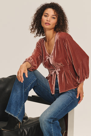 A woman sits on a chair wearing the ELIDA TOP by Velvet by Graham & Spencer, featuring wide sleeves and pleated details, paired with blue jeans. She looks at the camera with a neutral expression for a chic, understated holiday look.