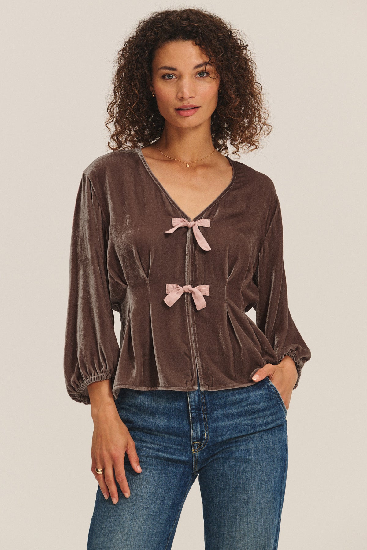 A woman wears the ELIDA TOP by Velvet by Graham & Spencer—a brown silk velvet blouse with pink ribbon bows—paired with blue jeans, standing against a plain light background for a charming holiday look.