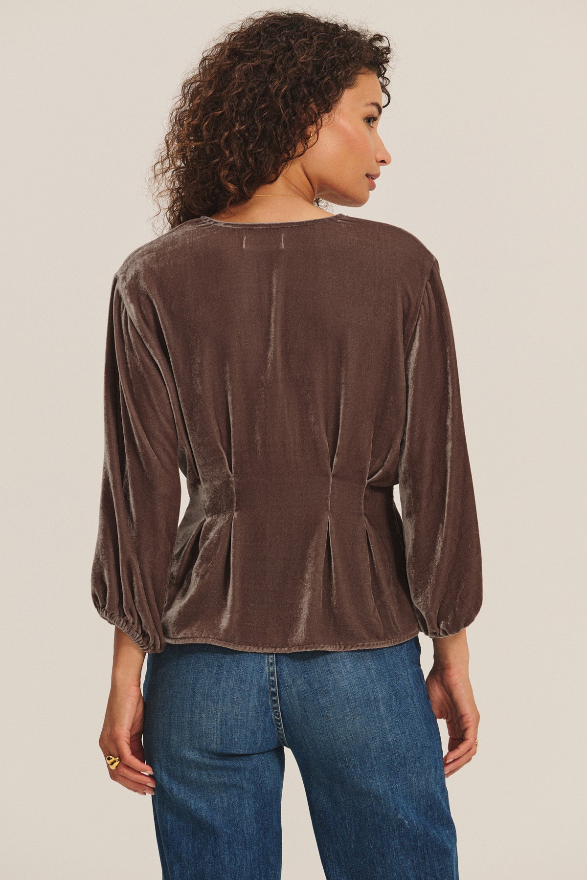 A woman with curly hair is shown from the back, wearing the ELIDA TOP by Velvet by Graham & Spencer in brown silk velvet and blue jeans, creating a festive holiday look against a plain light background.-40946746065089