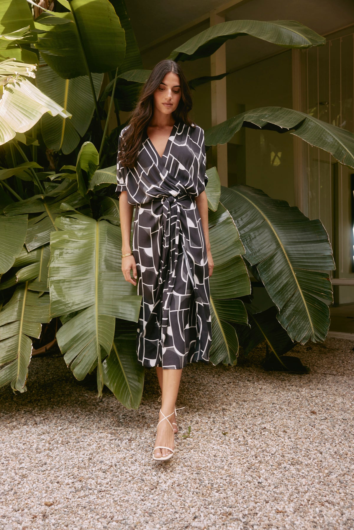   A woman wearing the ELISE DRESS, a stylish black and white piece by Velvet by Graham & Spencer featuring a Bauhaus-inspired print and V-neckline, strolls on gravel near large green leaves. 