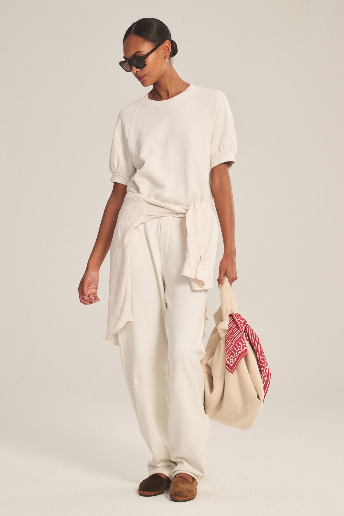 A woman wears the ELISE SWEATPANT by Velvet by Graham & Spencer as part of an all-white casual outfit with a sweater tied at the waist, brown shoes, sunglasses, and a beige tote bag featuring red detail.-41038249722049