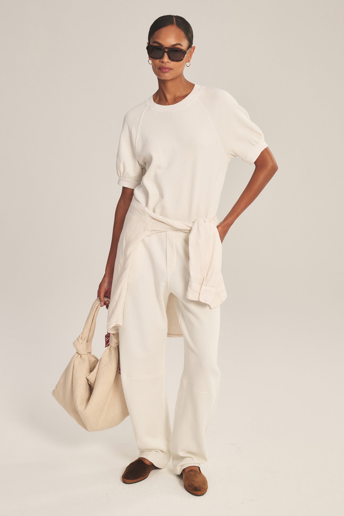 A woman wears the Velvet by Graham & Spencer ELISE SWEATPANT with a white short-sleeve top, brown shoes, cream sweater tied at her waist, beige bag in hand, and sunglasses.-41038249656513